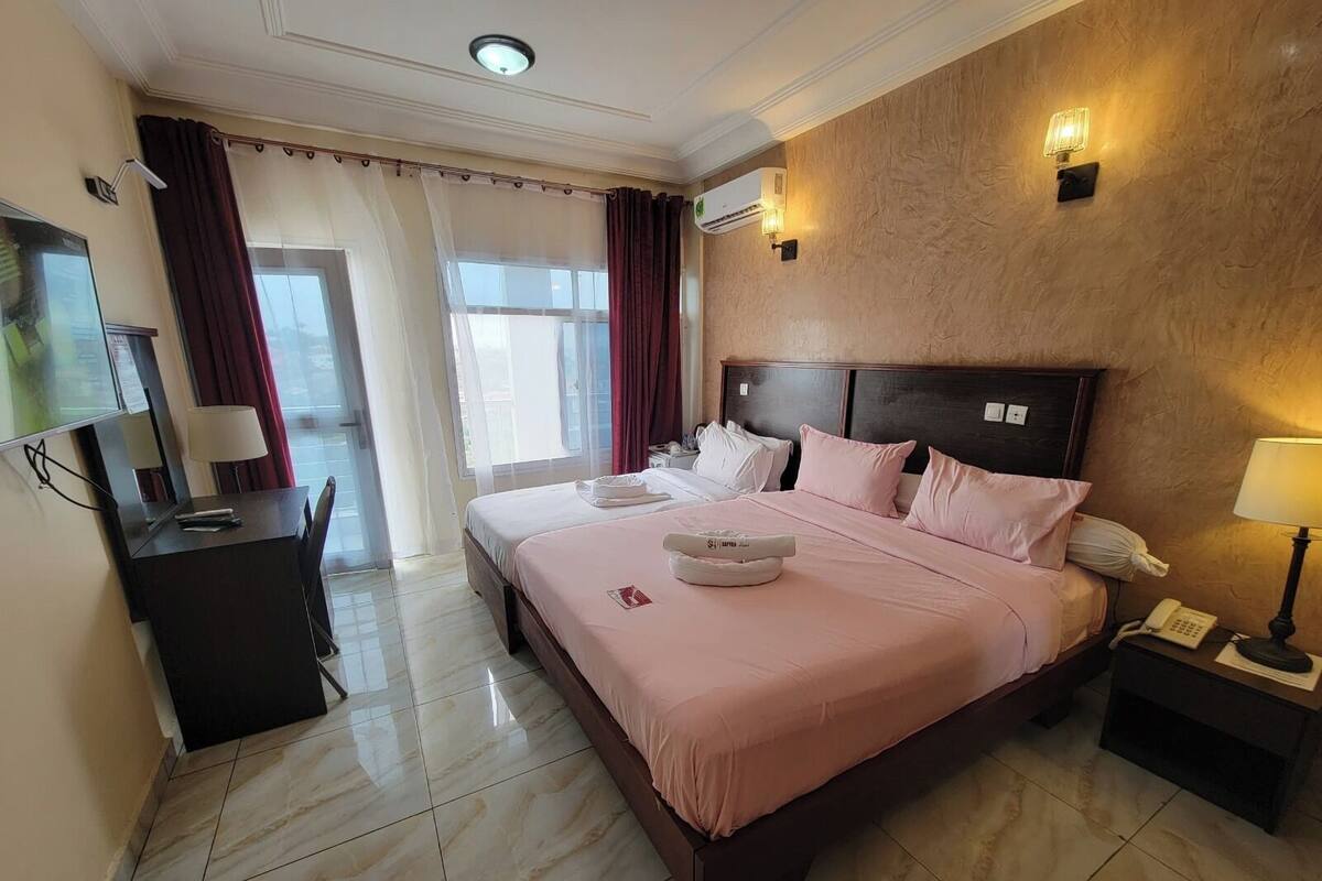 Quarto de hotel com cama, mesas de cabeceira, escrivaninha e televisão.