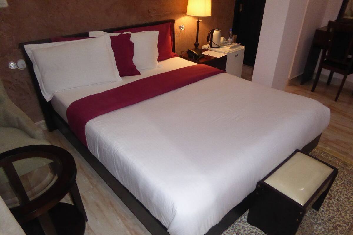 Quarto de hotel com uma cama grande, uma mesinha de cabeceira com abajur, uma cadeira e uma pequena mesa.