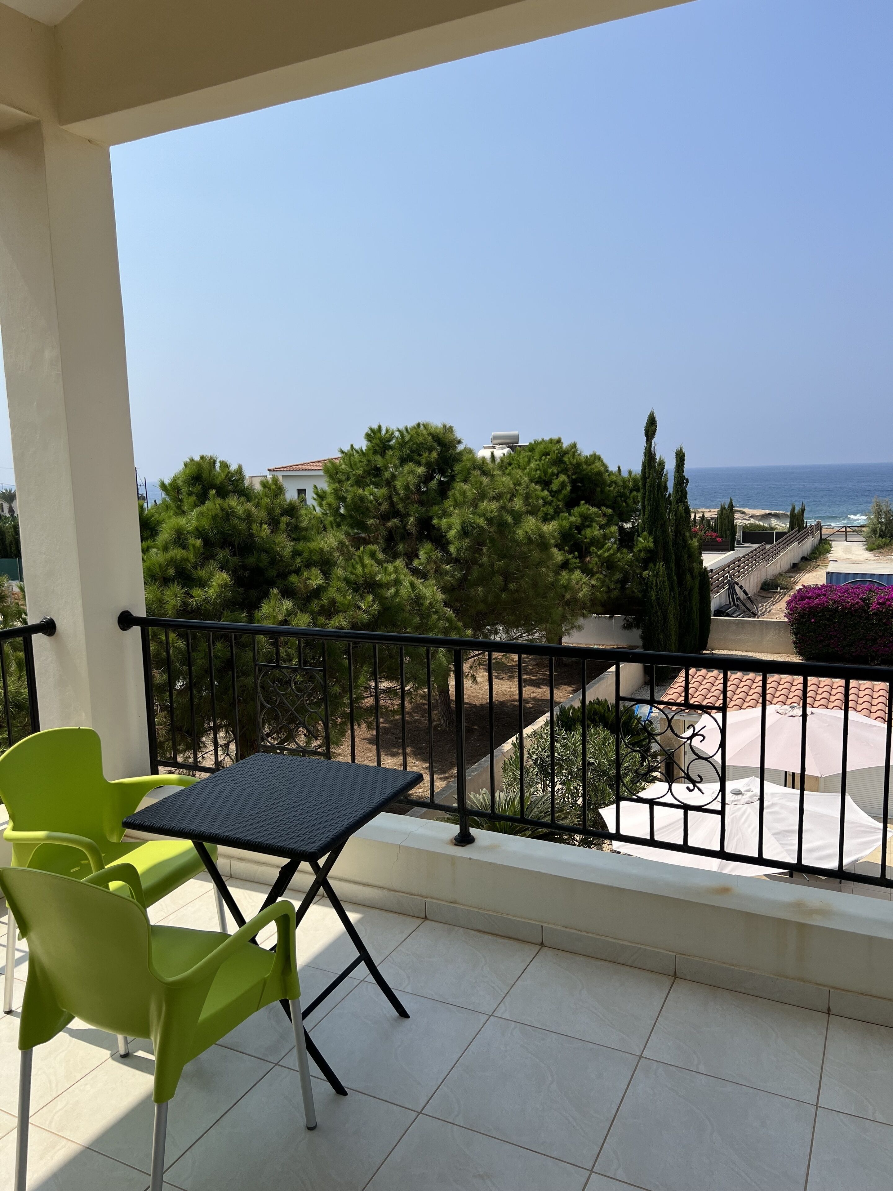 A balcony with a table and chairs, overlooking a beach and a garden with cypress trees.