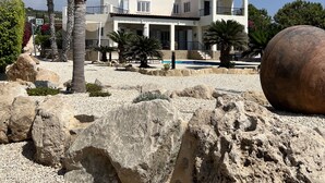 A modern two-story house with a balcony, surrounded by palm trees and large rocks. - Villa With Private Pool And Stunning Sea Views And Large Landscaped Gardens (Peyia)