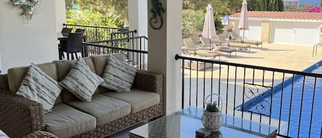 Un patio iluminado por el sol con mobiliario de mimbre, una mesa de centro de vidrio y vistas a una piscina y follaje.