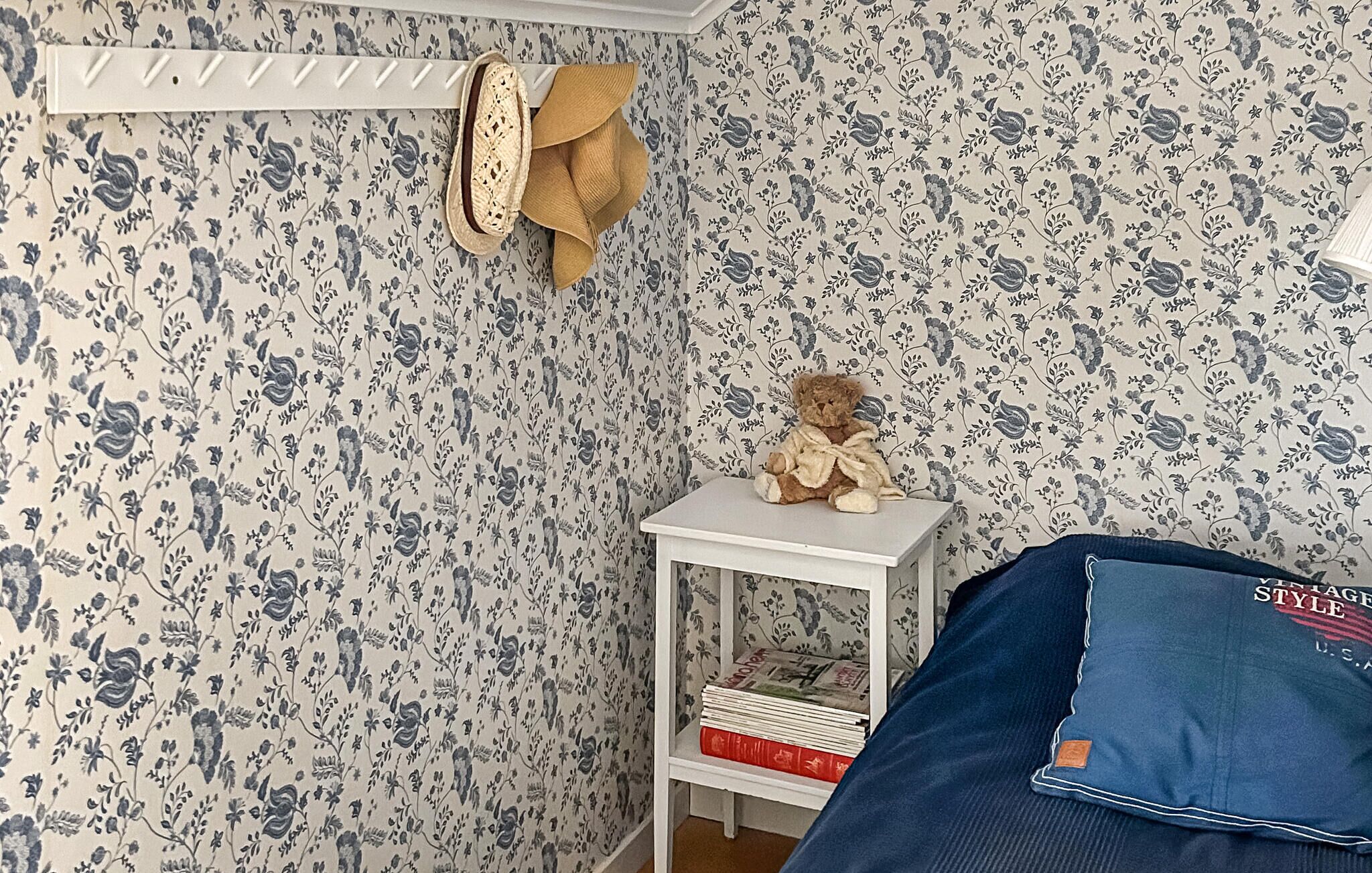 A room with floral wallpaper, a white shelf with books, a teddy bear, and a blue pillow.