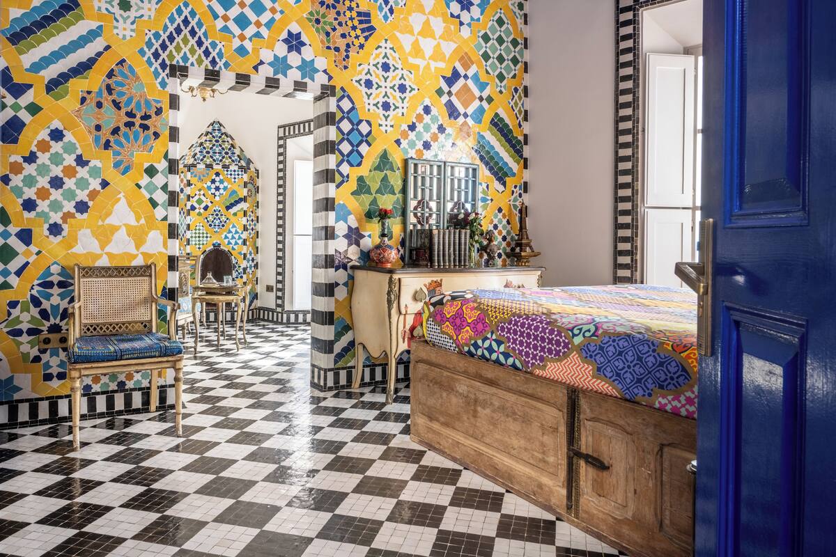 Une pièce avec un sol à carreaux, un lit recouvert d’une couverture aux couleurs vives, une chaise en bois et un mur orné d’un motif géométrique en carreaux.