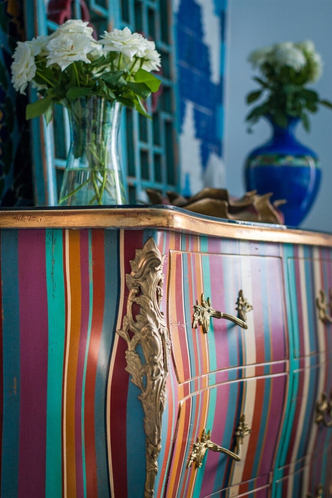 A colorful striped cabinet with ornate handles and a vase of white flowers on top.
