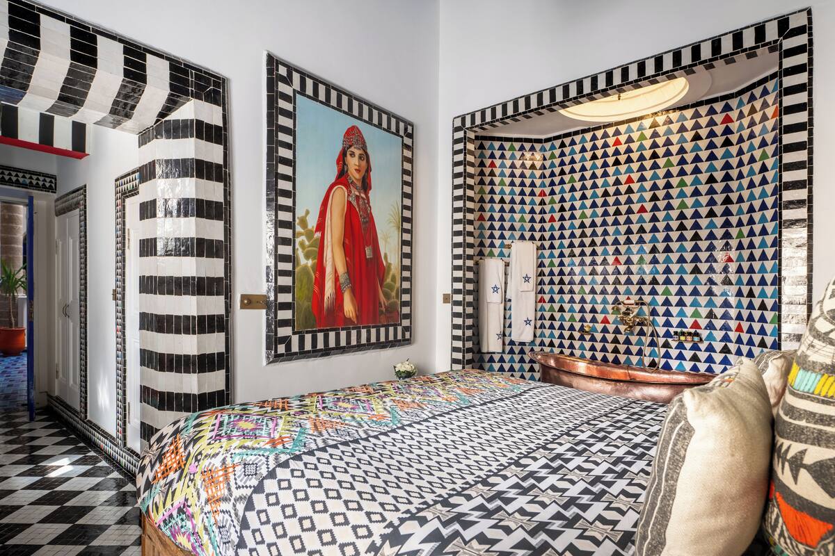 Une chambre à coucher avec une couvre-lit à motifs, un grand tableau encadré, un mur recouvert de carreaux formant un motif géométrique et une tête de lit originale.