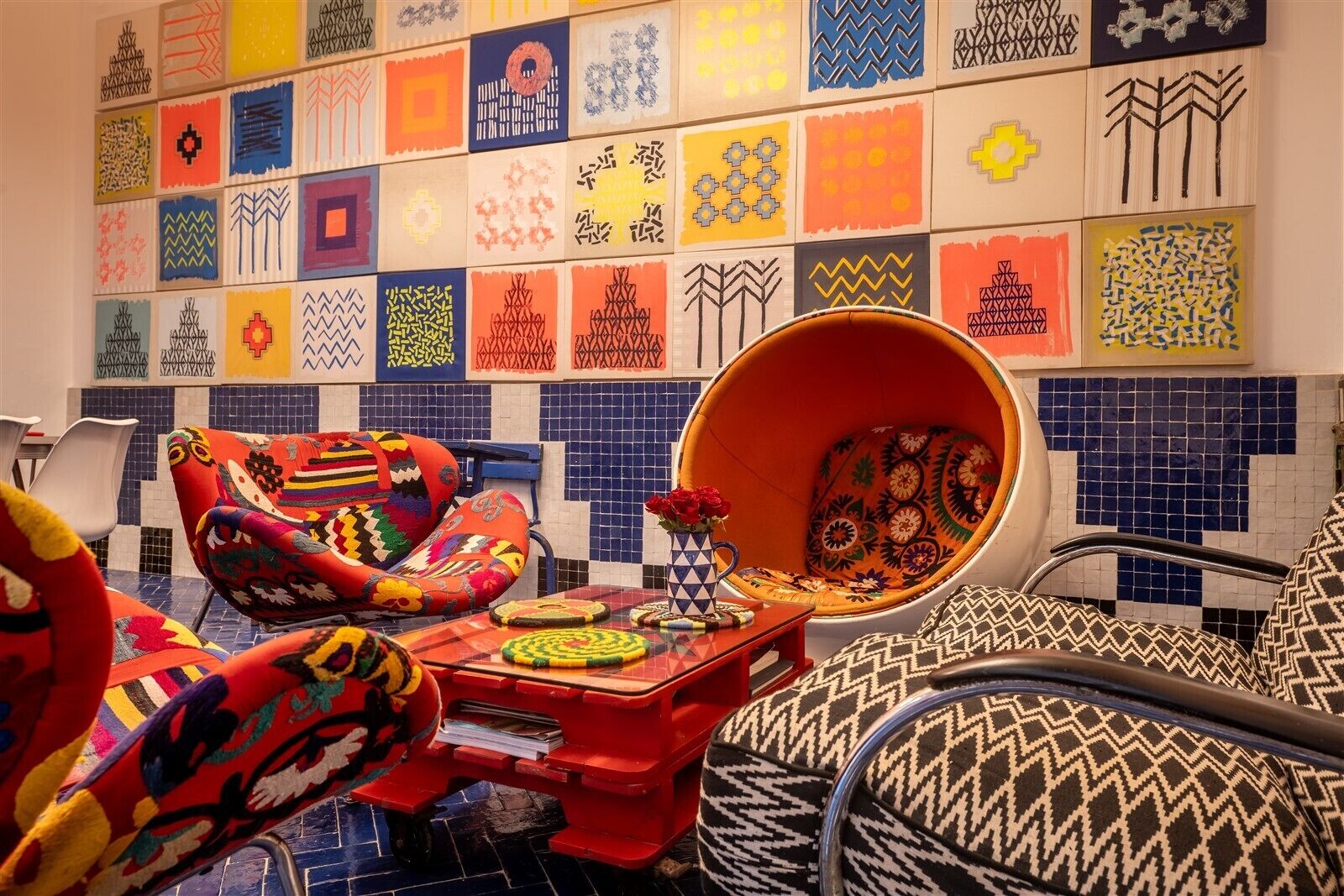Una habitación vibrante con almohadones de colores y diseños estampados, un sillón redondo naranja y una pared de azulejos con diversos diseños geométricos.