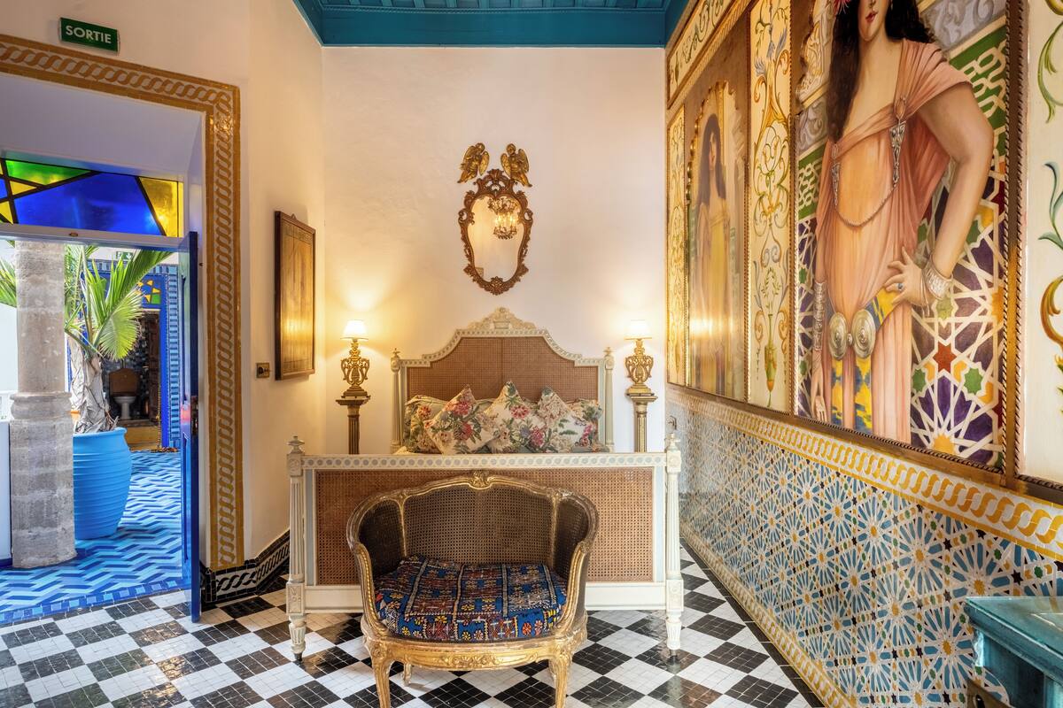 Une pièce avec un sol à carreaux, une chaise en rotin, un lit agrémenté d’oreillers à motifs floraux et une grande fresque murale.