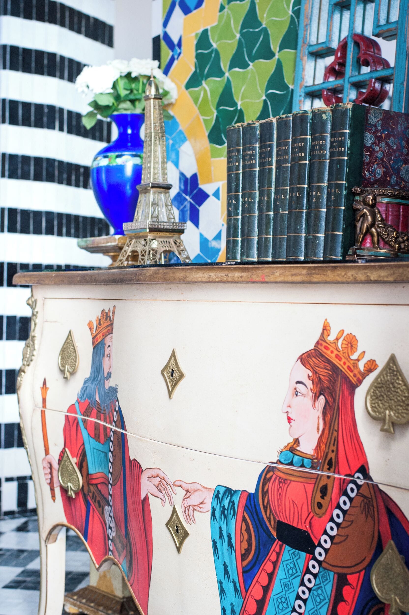 Un meuble décoratif orné d’une figure royale peinte, un vase bleu rempli de fleurs et une pile de livres.