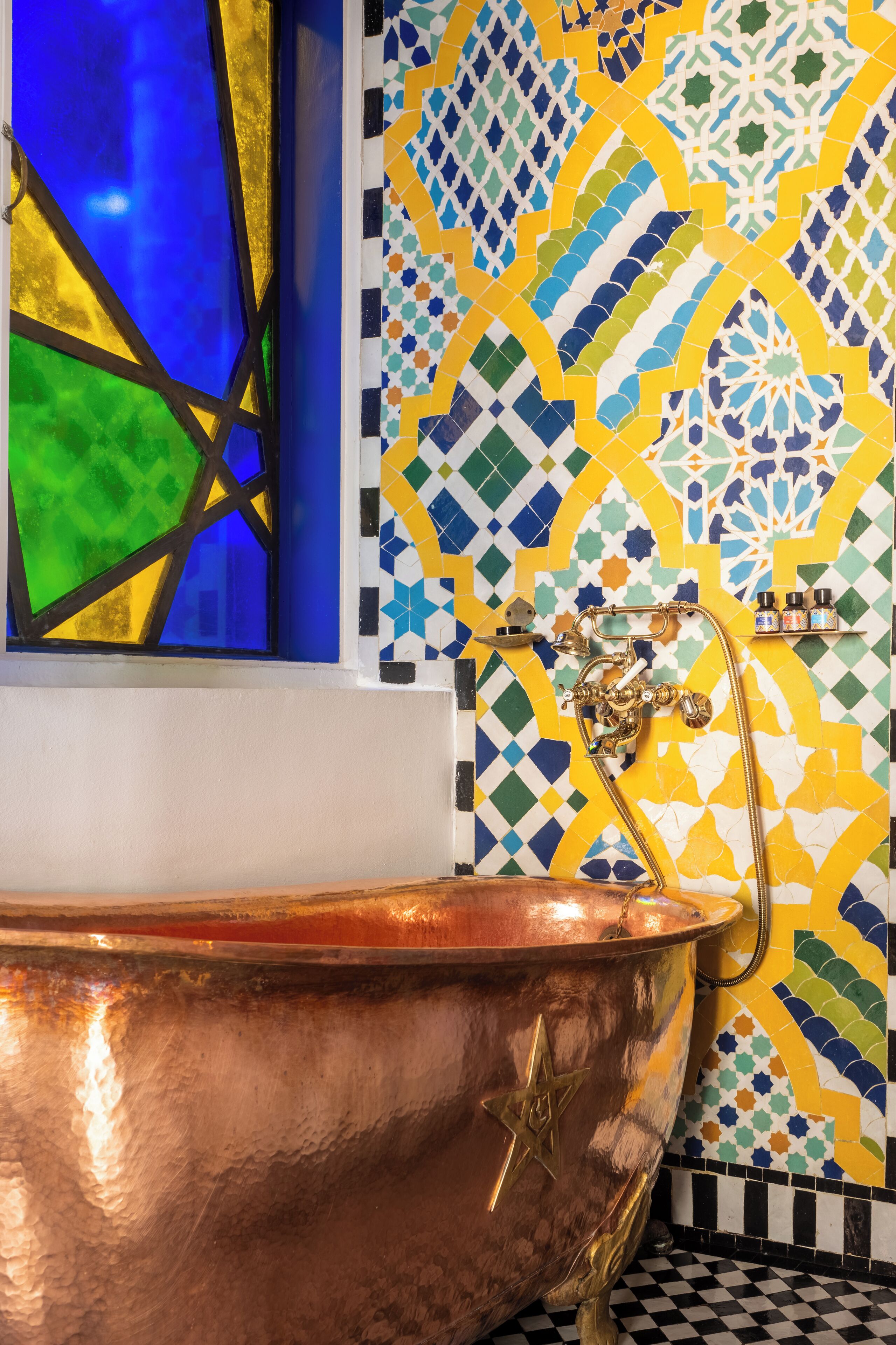 A copper bathtub with a star emblem, a window with blue and green glass, and a tiled wall with geometric patterns.