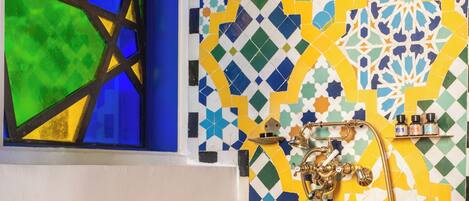 A copper bathtub with a star emblem, a window with blue and green glass, and a tiled wall with geometric patterns.