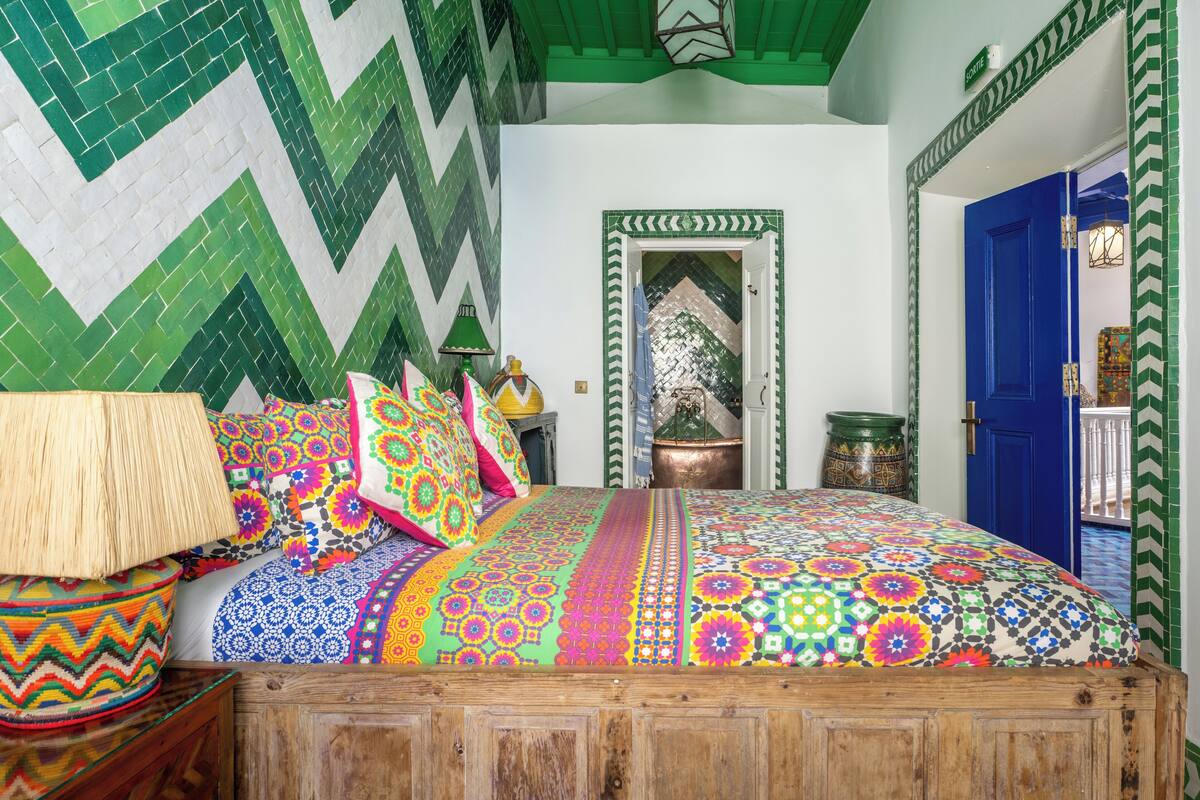 Une chambre avec un lit, des coussins colorés, une lampe et un mur orné d’un motif vert et blanc.