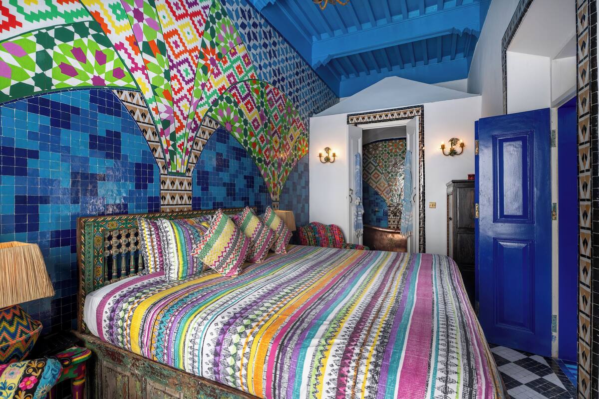 Une pièce avec un plafond orné d’une mosaïque colorée, un grand lit avec une literie à motifs et une porte bleue.