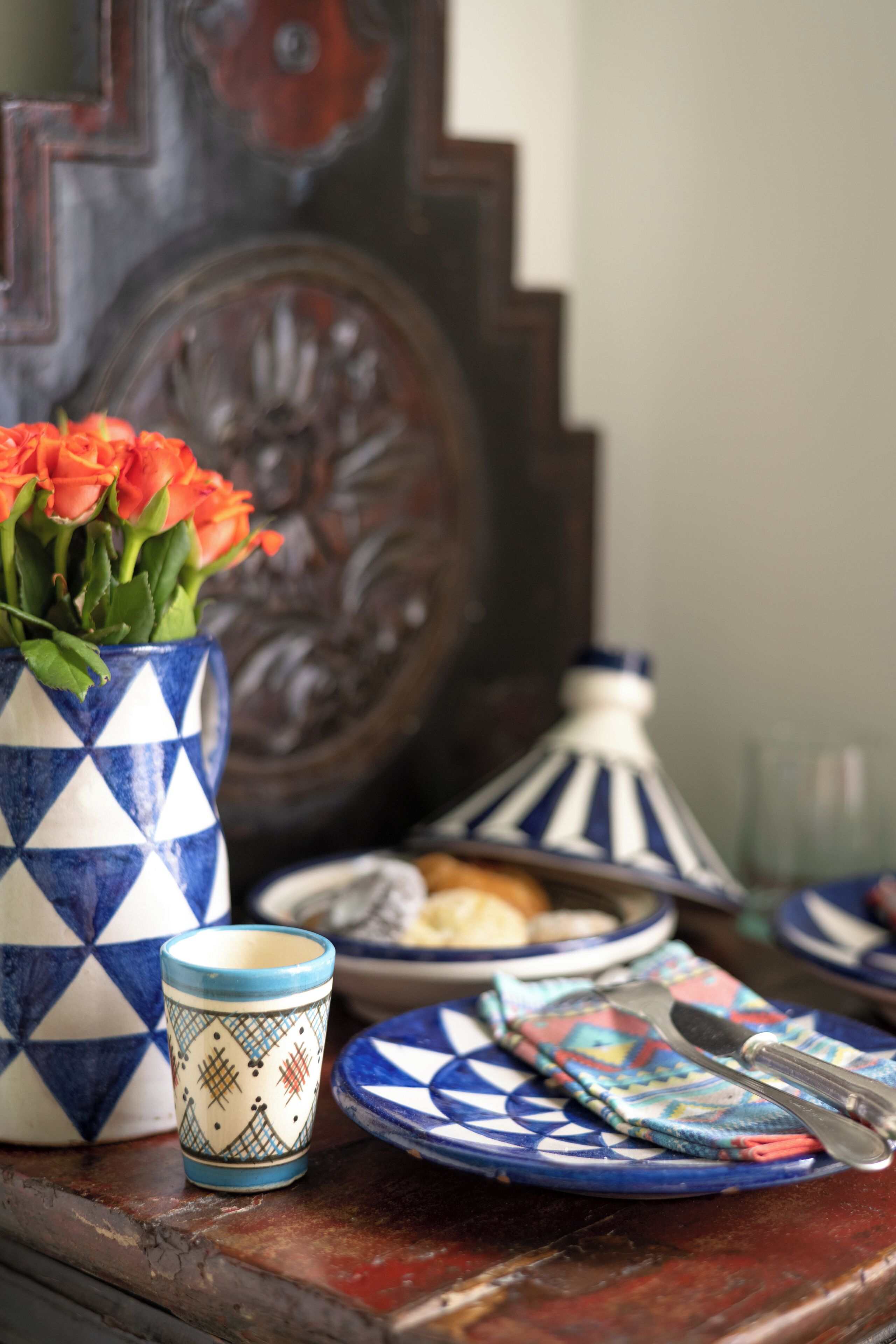 Una mesa con un jarrón azul y blanco de estampado, flores naranjas, una taza pequeña y un plato con tenedor y cuchillo.