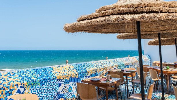 Un restaurant en bord de mer avec une terrasse, un sol à motifs et un parasol en chaume.