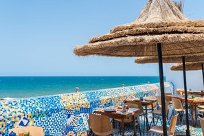 Un restaurant en bord de mer avec une terrasse, un sol à motifs et un parasol en chaume.