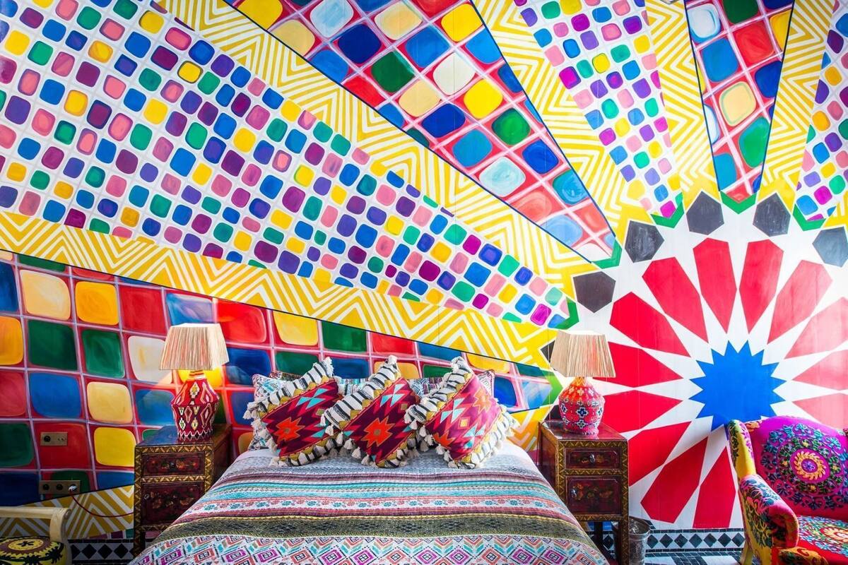 Une pièce avec un mur décoré d’une mosaïque colorée, un lit avec une literie à motifs et un fauteuil décoratif.