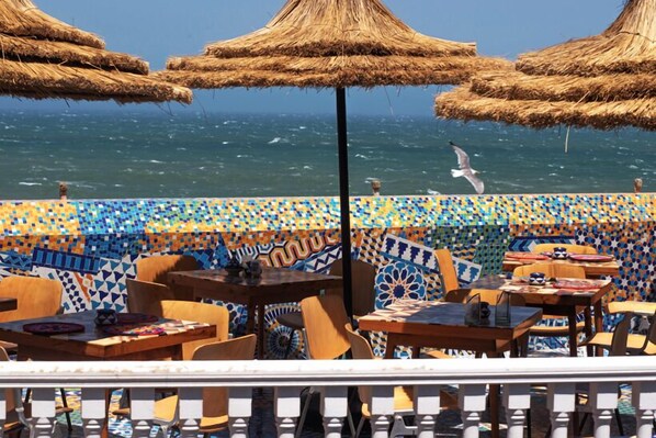 Un café situé en bord de mer, doté de parasols en chaume, de murs recouverts de carreaux colorés et de mobilier en bois.