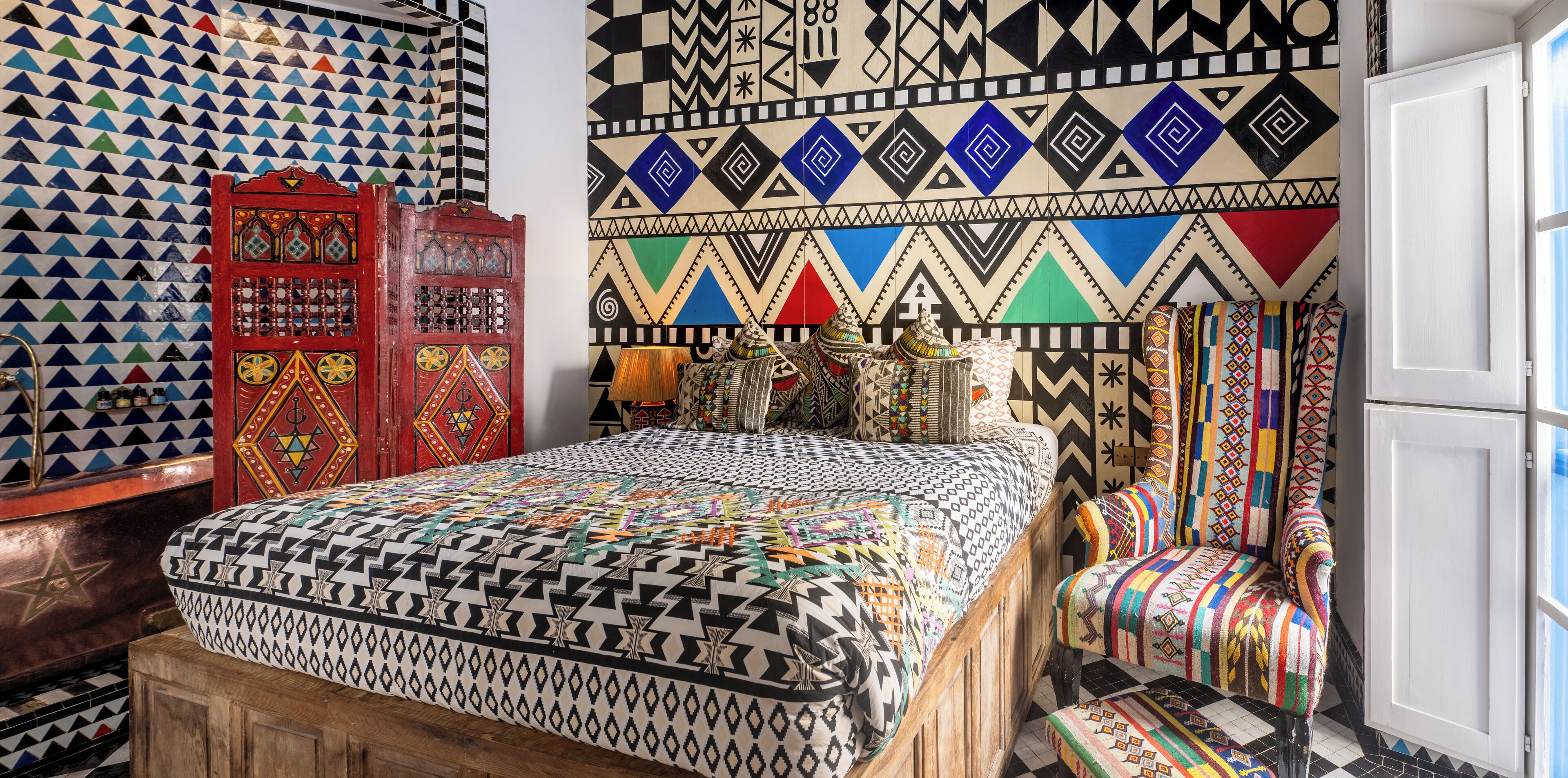 A room with a patterned bedspread, a red cabinet, and a wall with geometric designs.