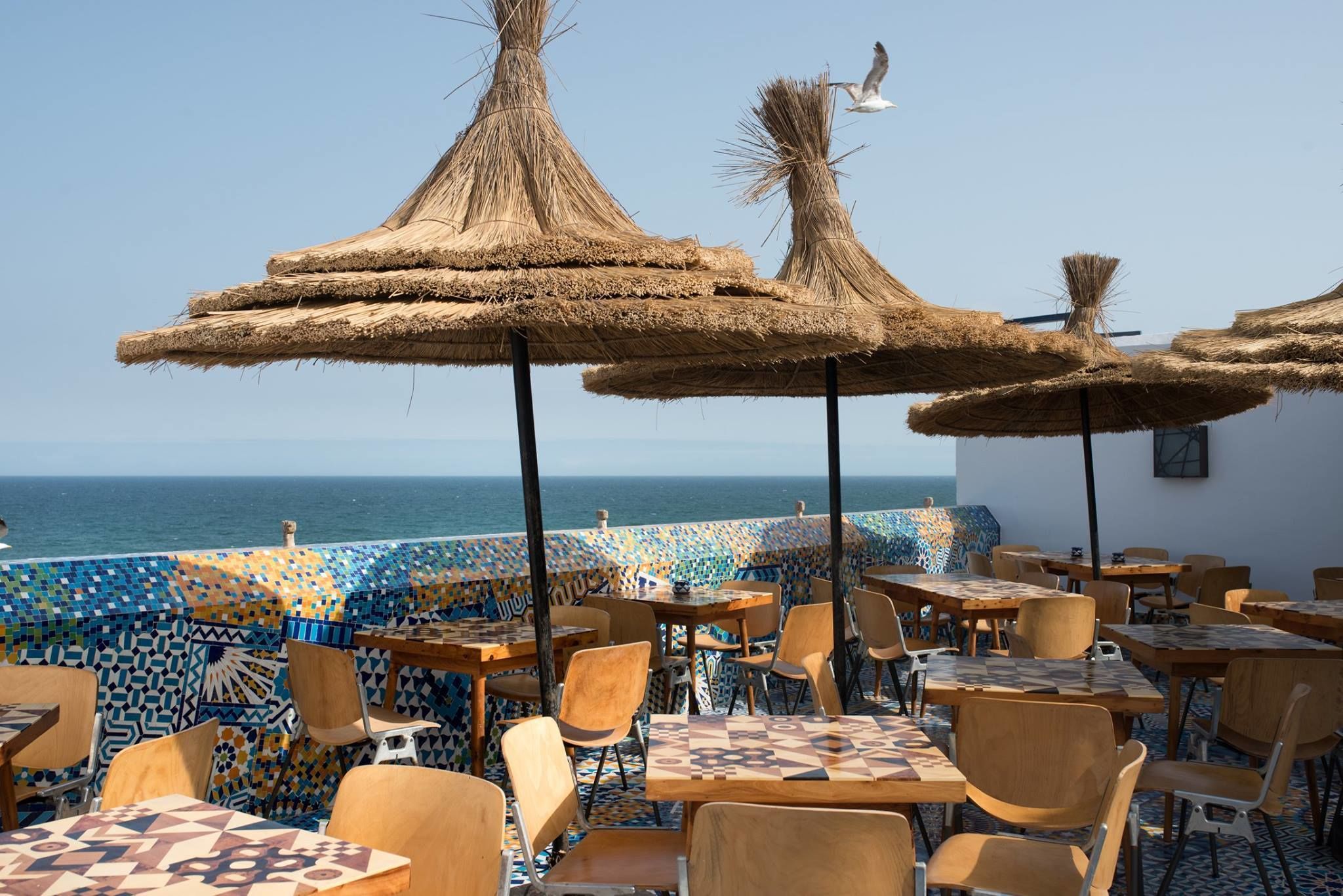 An outdoor seating area with tables and chairs, thatched umbrellas, and a view of the sea.
