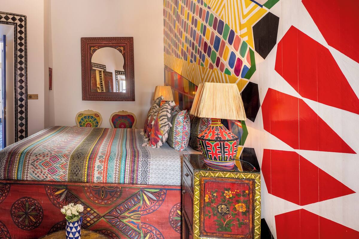 Une pièce avec un sol à carreaux, un lit recouvert d’une couverture colorée, un miroir et un mur orné de motifs géométriques.