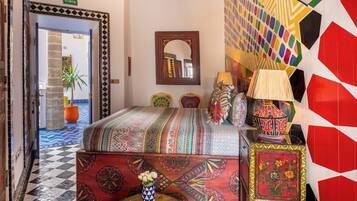 A room with a checkered floor, a bed with a colorful blanket, a mirror, and a wall with geometric patterns.