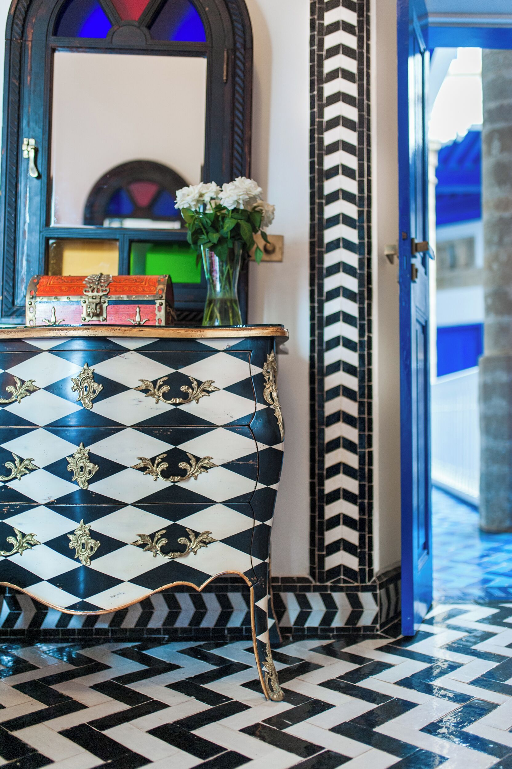 A checkered floor, a mirror, a vase with flowers, and a chest of drawers.
