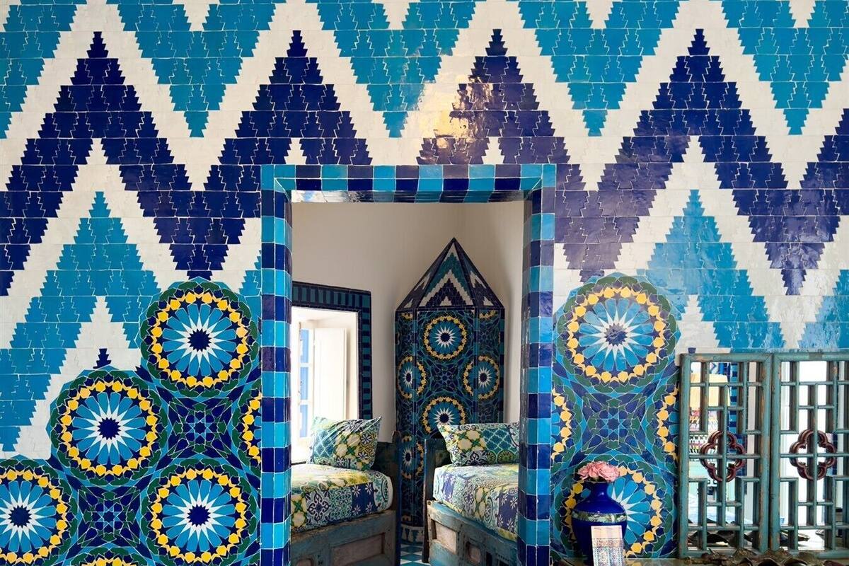 Une pièce dont un mur est orné d’un motif géométrique, dont le sol est à carreaux et qui contient un lit recouvert d’une couvre-lit à motifs.