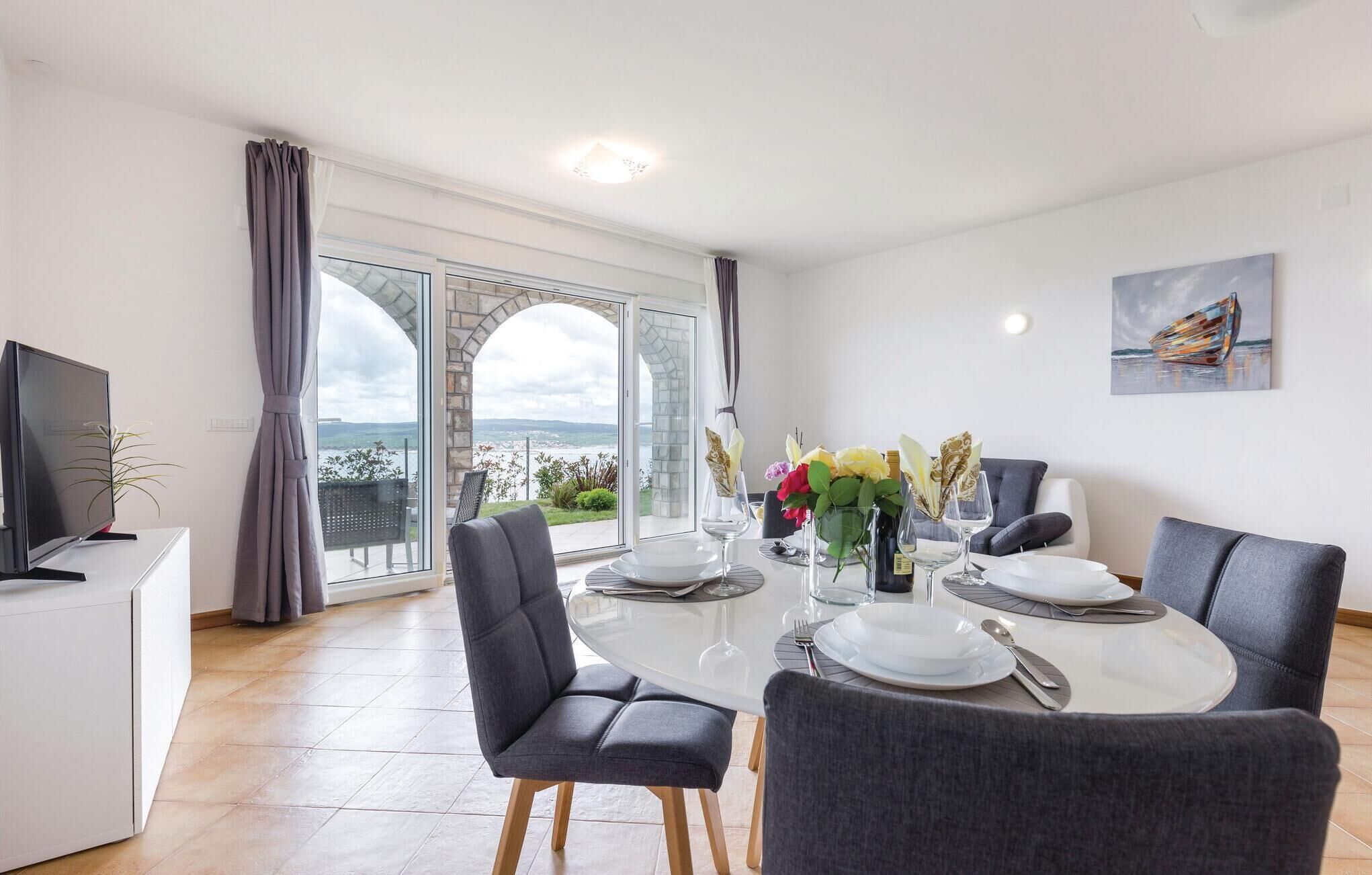 A dining area with a glass table, grey upholstered chairs, and a view of the sea through large windows.
