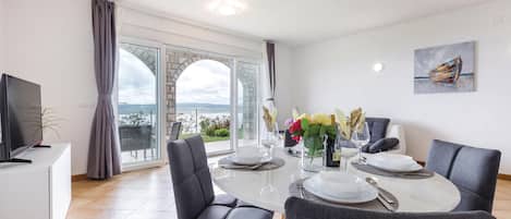 Un coin repas avec une table en verre, des chaises à revêtement gris et une vue sur la mer grâce à de grandes fenêtres.