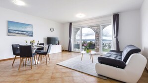 Un soggiorno moderno con divano, tavolo da pranzo, sedie, televisione e vista sul mare attraverso ampie finestre.