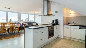 Une cuisine moderne avec des armoires blanches, une hotte de cuisinière en acier inoxydable et un four encastré. On y trouve un coin repas avec une table et des chaises, ainsi qu’une grande fenêtre qui laisse entrer la lumière naturelle.