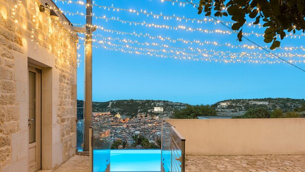 A stone building with a glass-enclosed pool, string lights, and a view of a town and hills. - Villa with pool and private wellness area in Scicli, Sicily - 8 pax (Scicli)