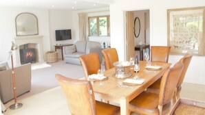 Un coin repas avec une table en bois dressée pour six personnes, entourée de fauteuils en cuir. On aperçoit une cheminée au fond, et un téléviseur est fixé au mur.