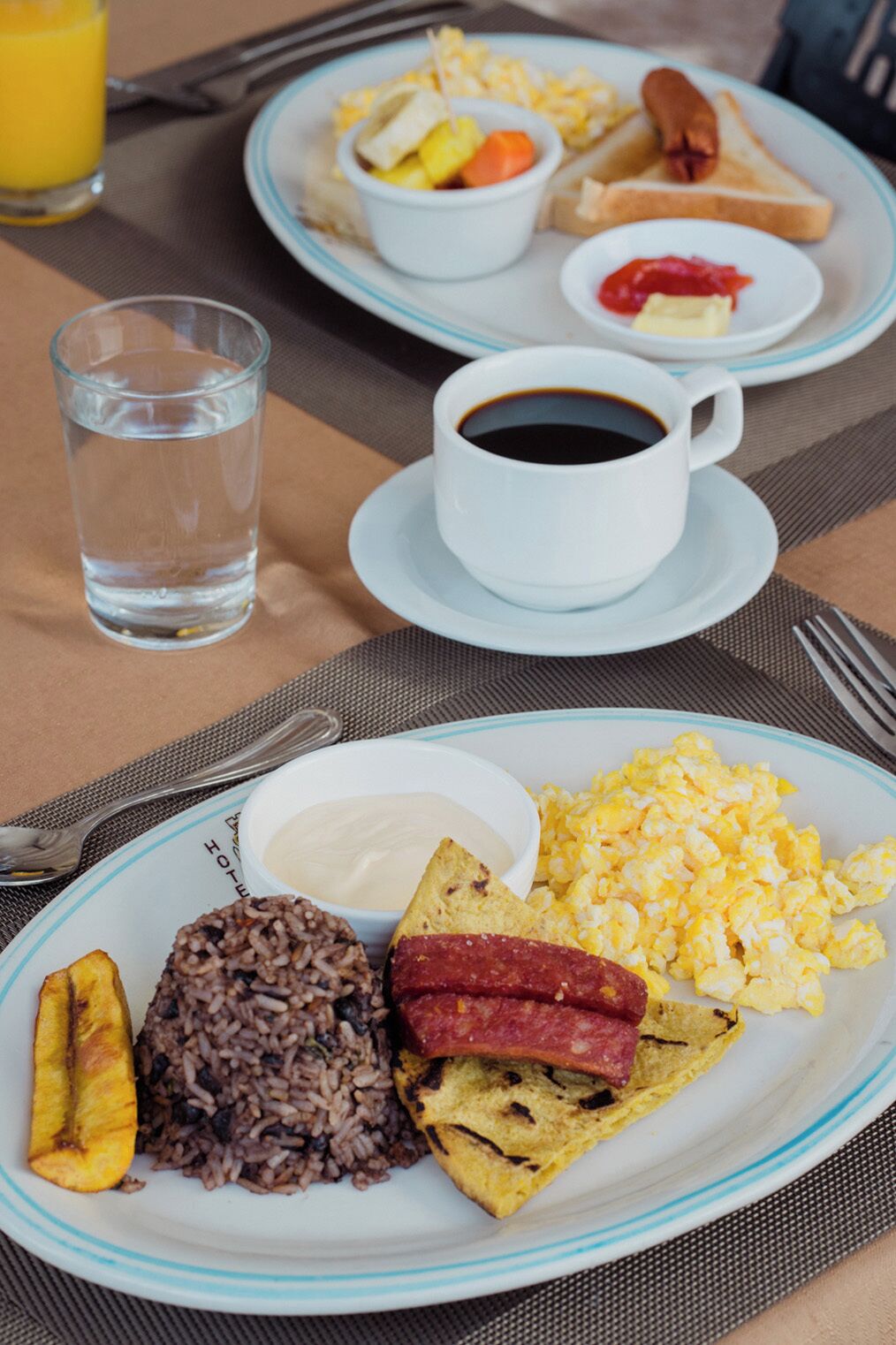 Une table dressée pour le petit-déjeuner avec des assiettes contenant des œufs brouillés, du riz noir, des bananes plantains frites et une tranche de jambon. On y trouve également des tasses de café et de jus de fruits.