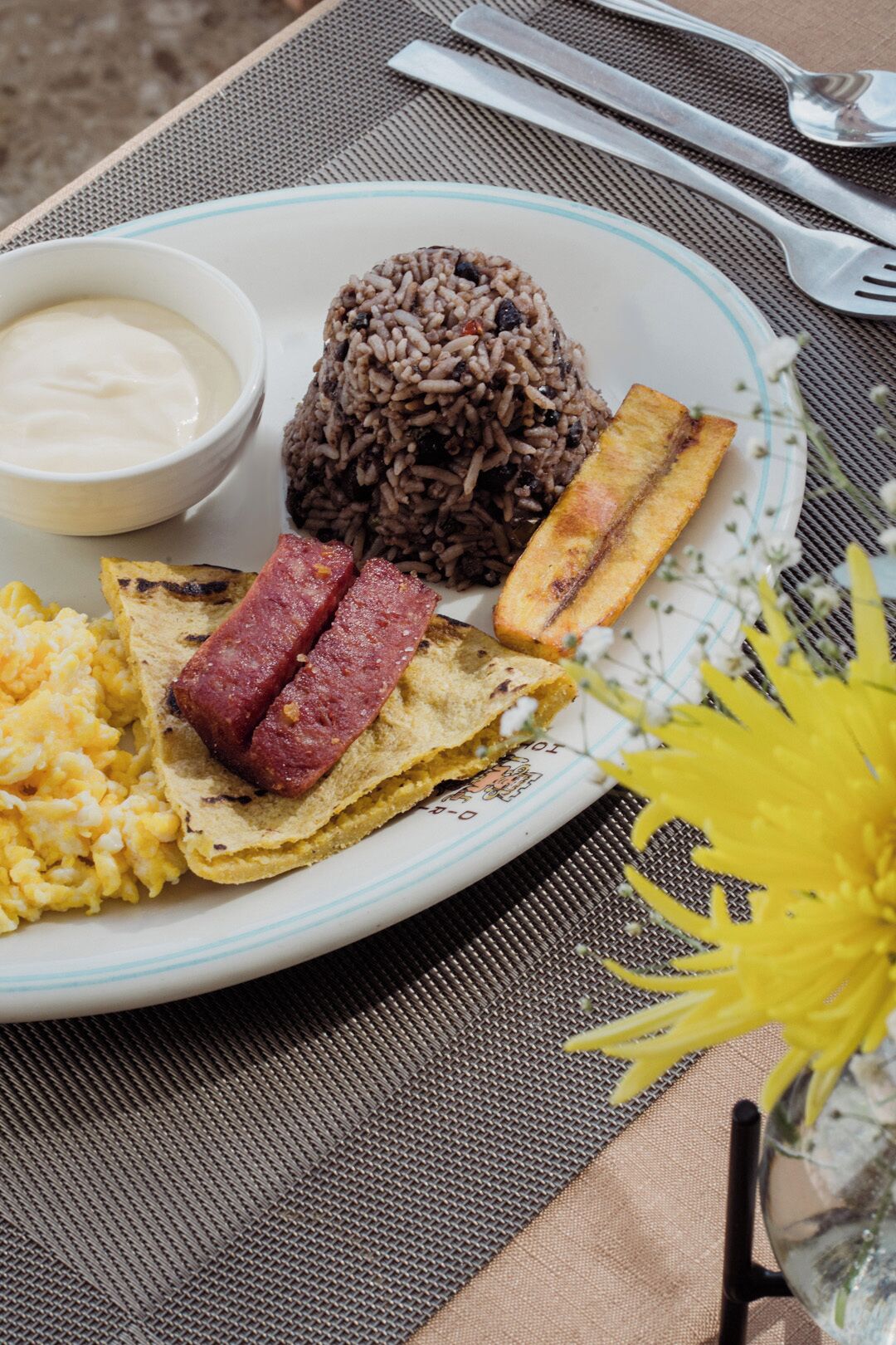 Un plato con arroz negro, plátanos fritos, huevos revueltos y una rebanada de tostada con salchicha.