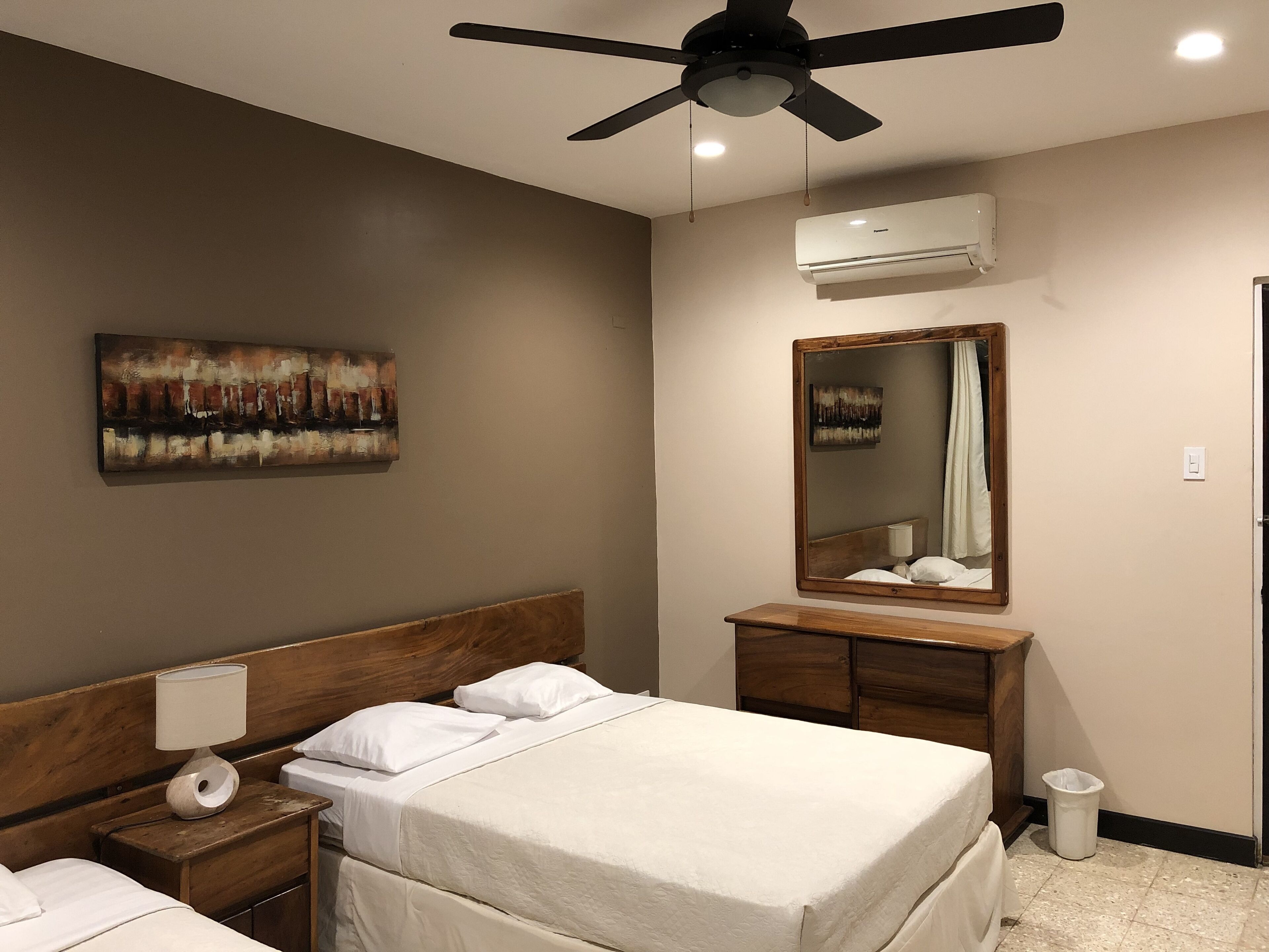 Une chambre d’hôtel avec un lit, une table de chevet, un miroir et un ventilateur de plafond.