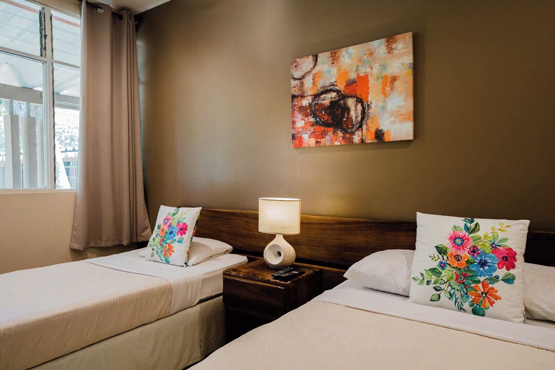 A hotel room with two beds, a nightstand with a lamp, a floral pillow, and a painting on the wall.