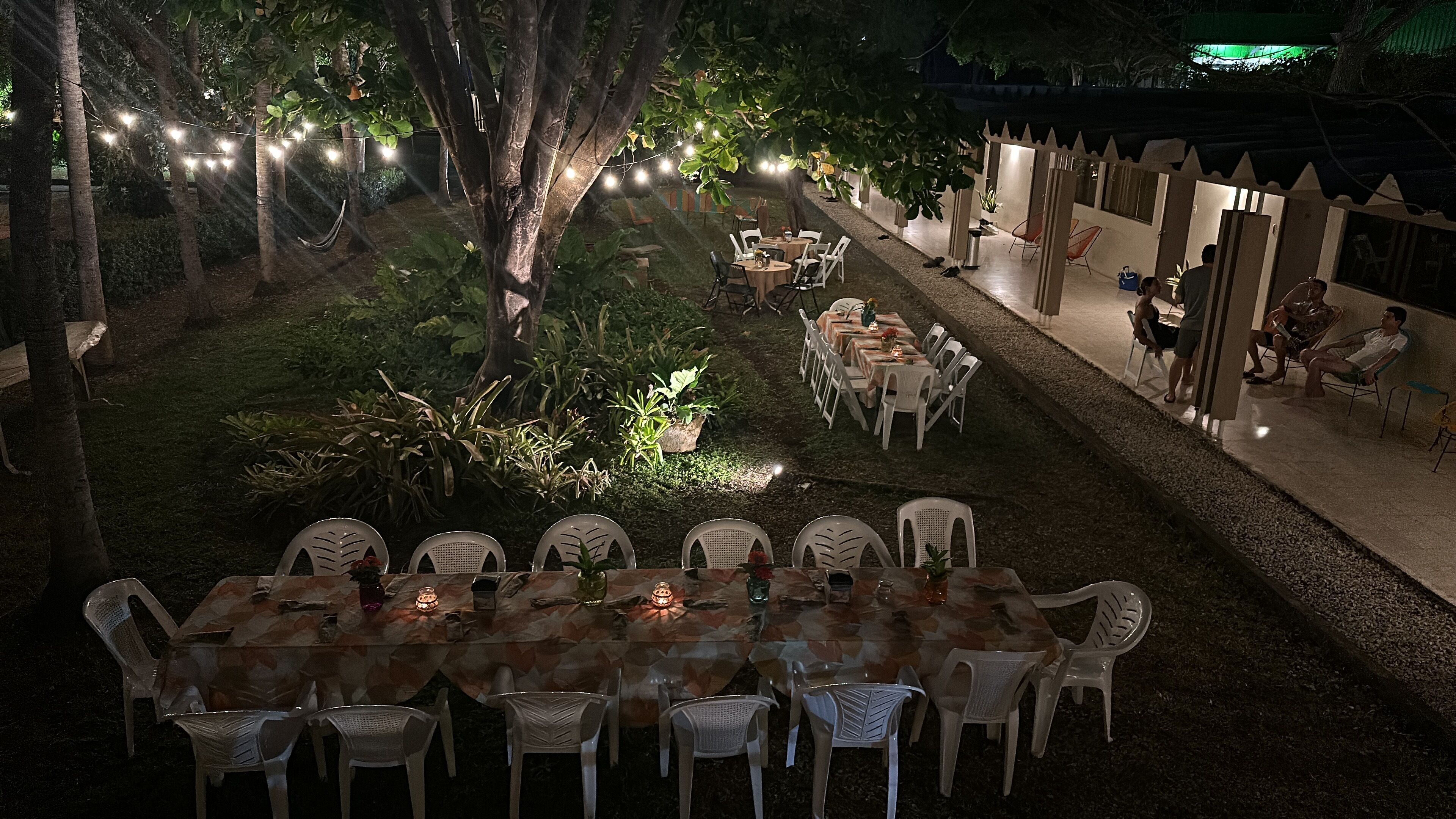 Un área de comedor al aire libre con mesas y sillas, rodeada de árboles y luces colgantes.
