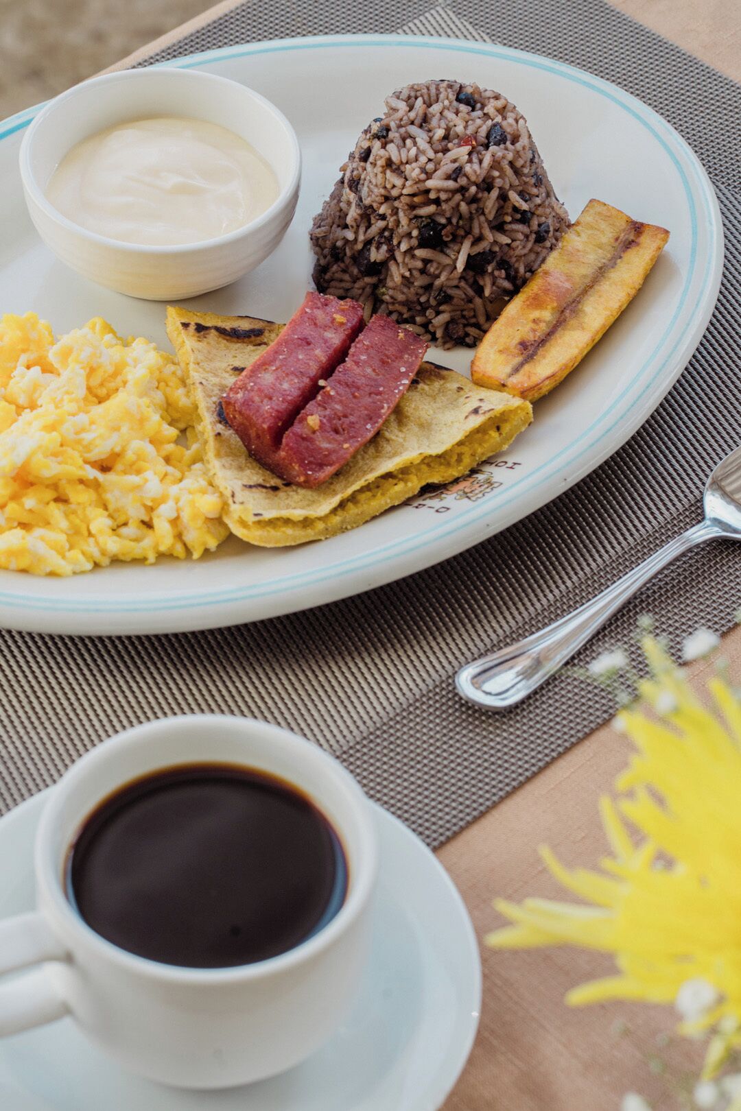 Um prato com arroz, ovos mexidos, presunto, banana frita e uma xícara de café preto.