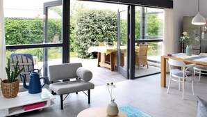 A modern living room with a large sliding glass door leading to a patio. There is a sofa, a coffee table, and a dining area with a table and chairs.