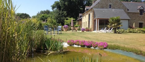 A stone building with a sloped roof, a pond with pink flowers, and outdoor seating.