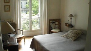 A bedroom with a bed, a chair, a desk with a lamp, a window with a view of greenery, and a wall-mounted lamp.
