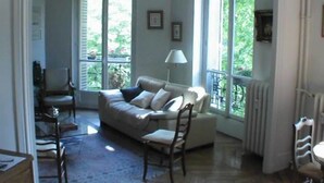 A cozy living room with a sofa, chairs, and a table. Large windows allow natural light and views of greenery outside.