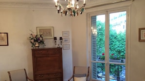 A room with a wooden cabinet, two upholstered chairs, a chandelier, and a large window with a view of greenery.