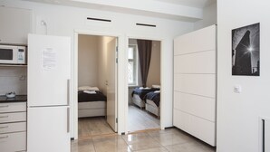 Eine kompakte Küche mit einer Mikrowelle, einem Kühlschrank und einer kleinen Arbeitsplatte. Ein Durchgang führt zu einem Schlafzimmer mit zwei Betten und einem Fenster.