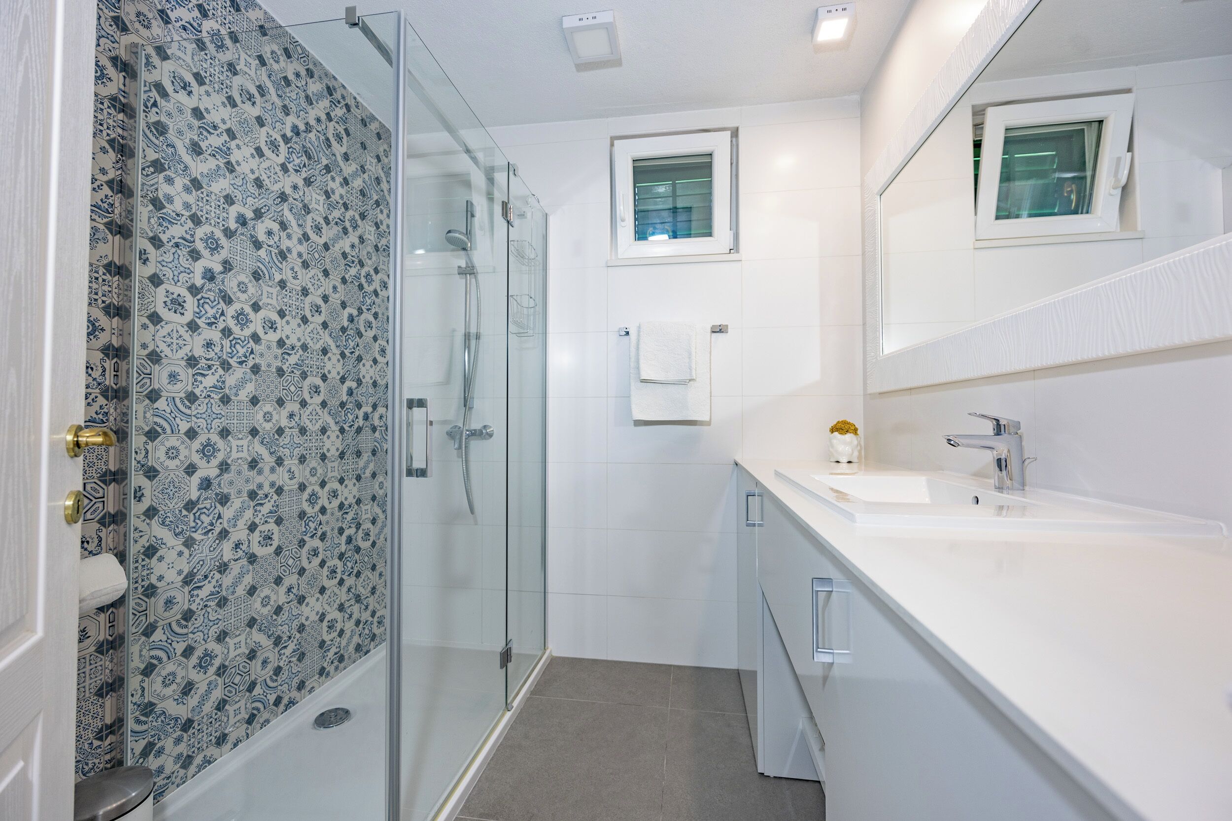 Une salle de bain moderne avec une douche à l’italienne en verre, un lavabo blanc et une petite fenêtre située au-dessus du miroir.