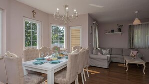 A dining area with a white table set for six, surrounded by cushioned chairs. There are two windows with curtains, a chandelier, and a grey sofa in the living area. - Amazing home in Imotski (Imotski)