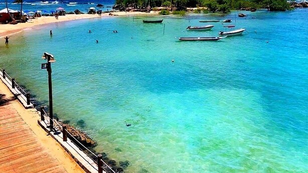 Una playa con agua turquesa cristalina, una pasarela de madera y varias embarcaciones ancladas cerca.