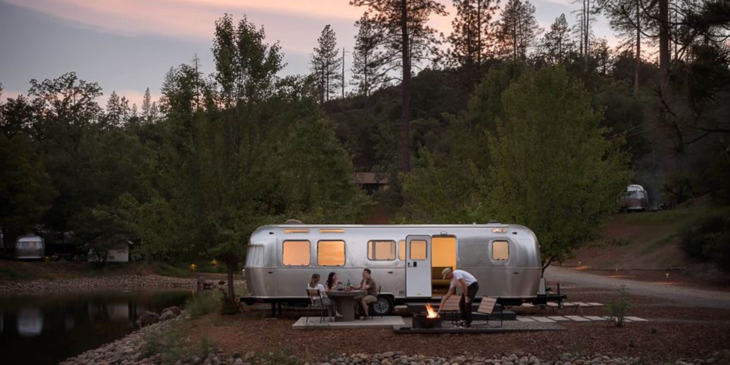A stationary RV with a fire pit in front, surrounded by trees and a body of water.