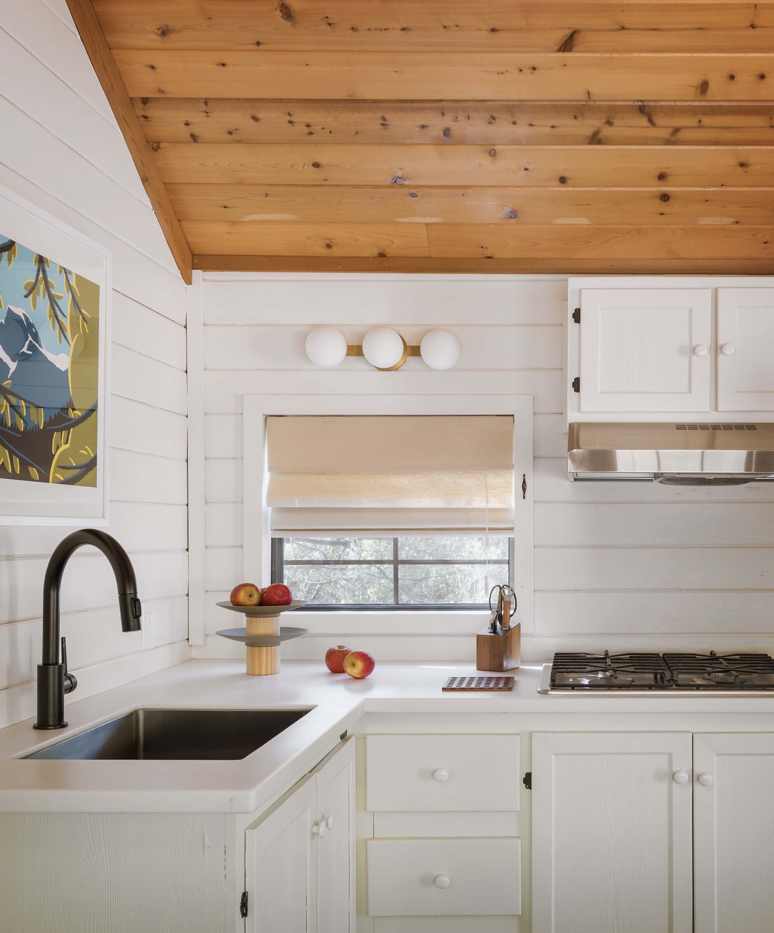 Cocina con muebles blancos, techo de madera, pileta de acero inoxidable y ventana con vista a árboles.